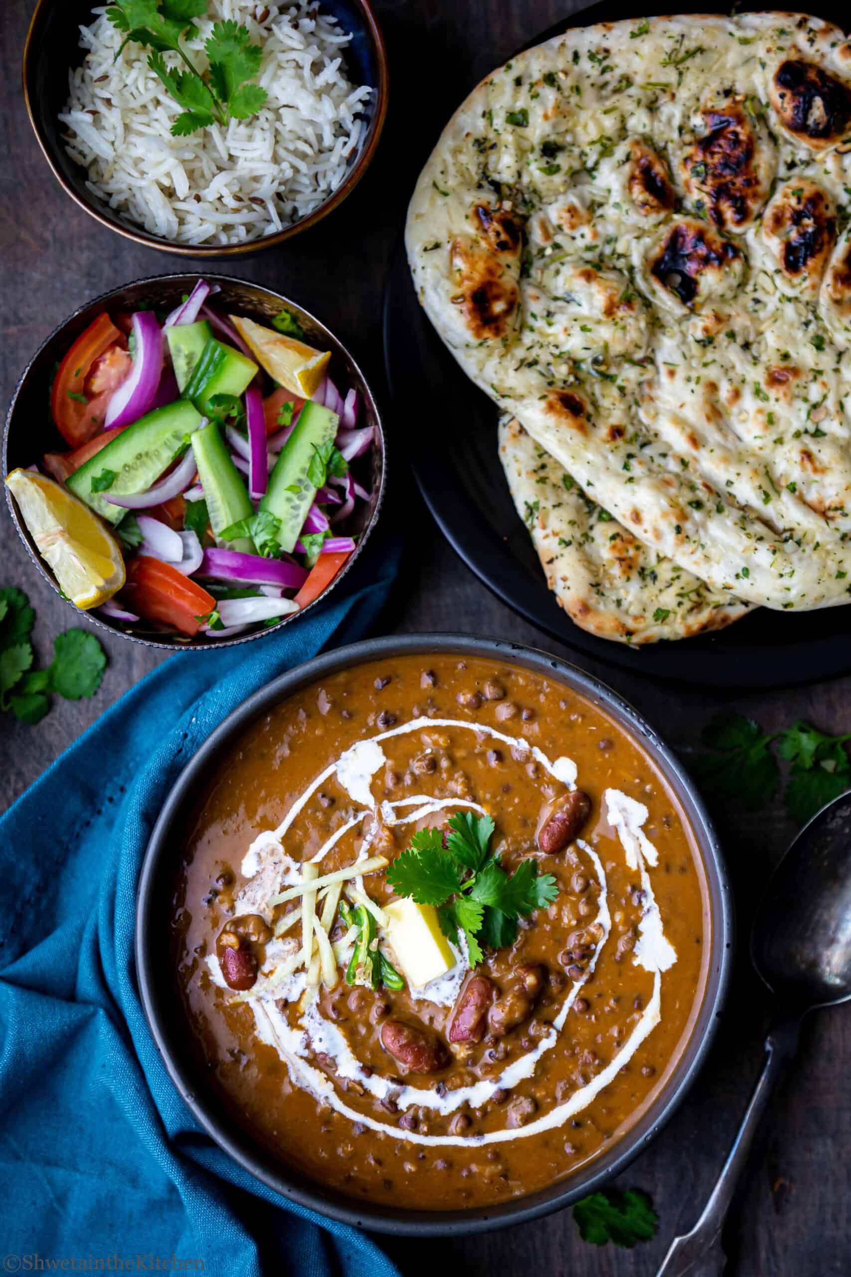 Dal Makhani in black bowl with another salad bowl, rice bowl and naan on side
