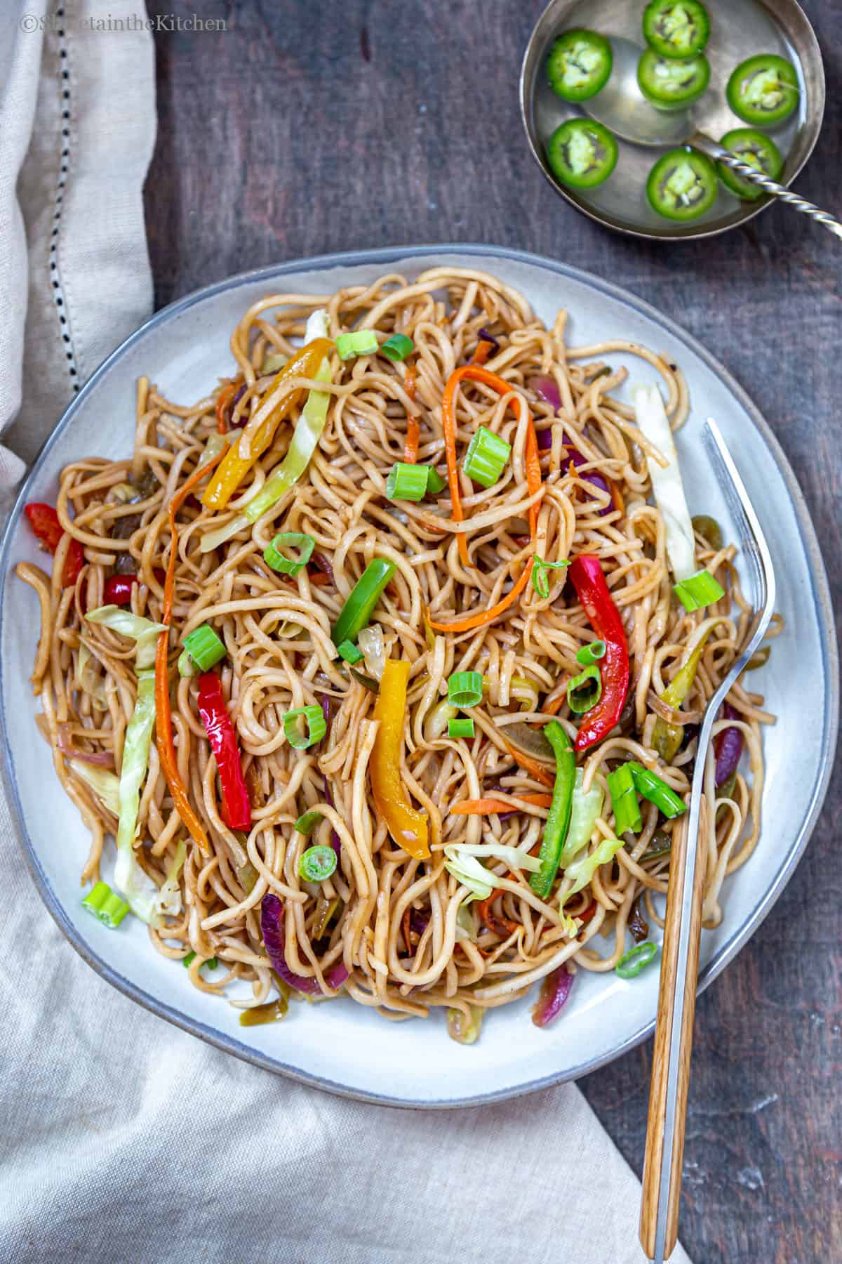 Vegetable hakka noodles on plate with fork on side and bowl of vinegar and chillies next to plate. 