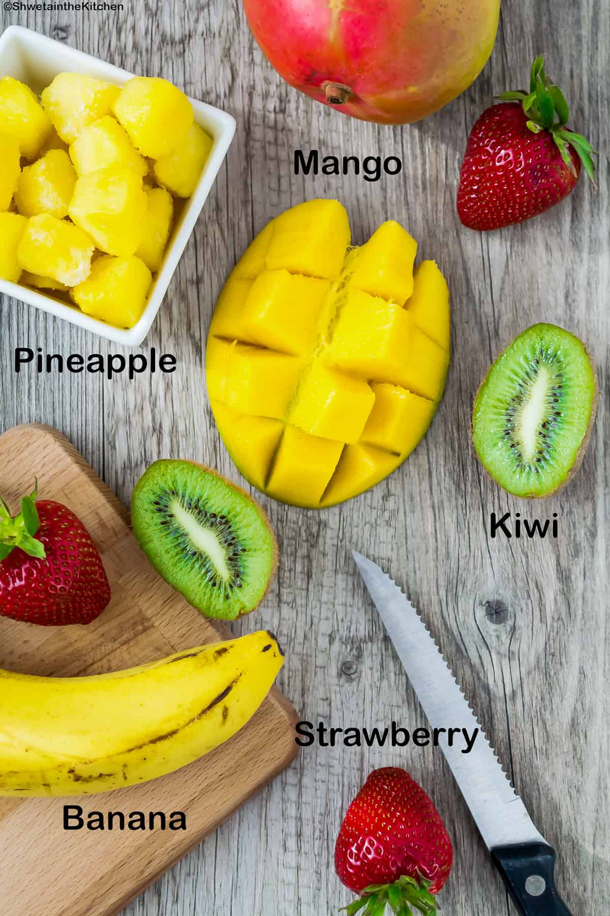Top view of cut mango, kiwi, few strawberries, bowl of pineapple pieces and banana on cutting board