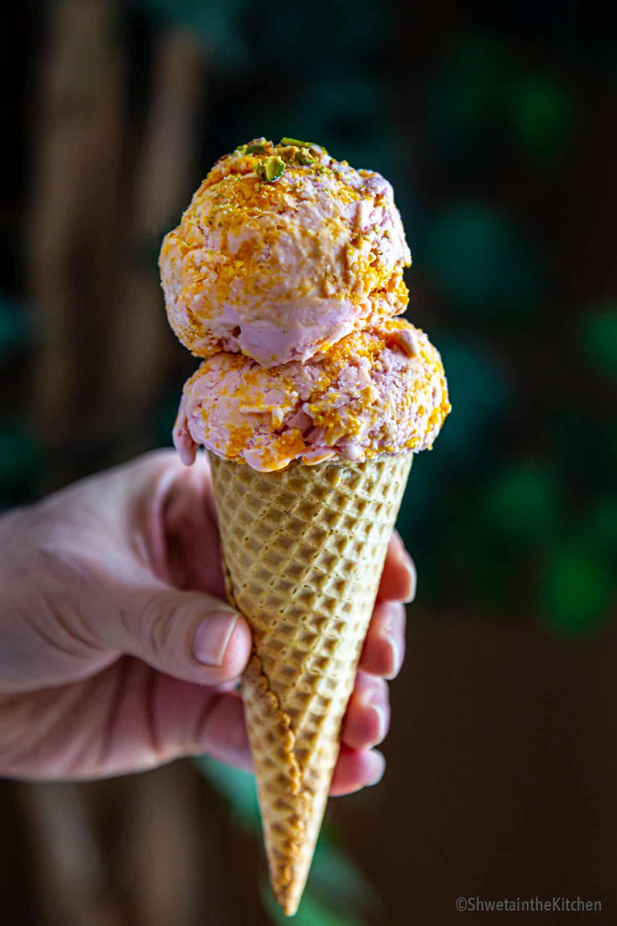 hand held waffle cone with two scoops of strawberry mango icecream garnished with pistachios. 