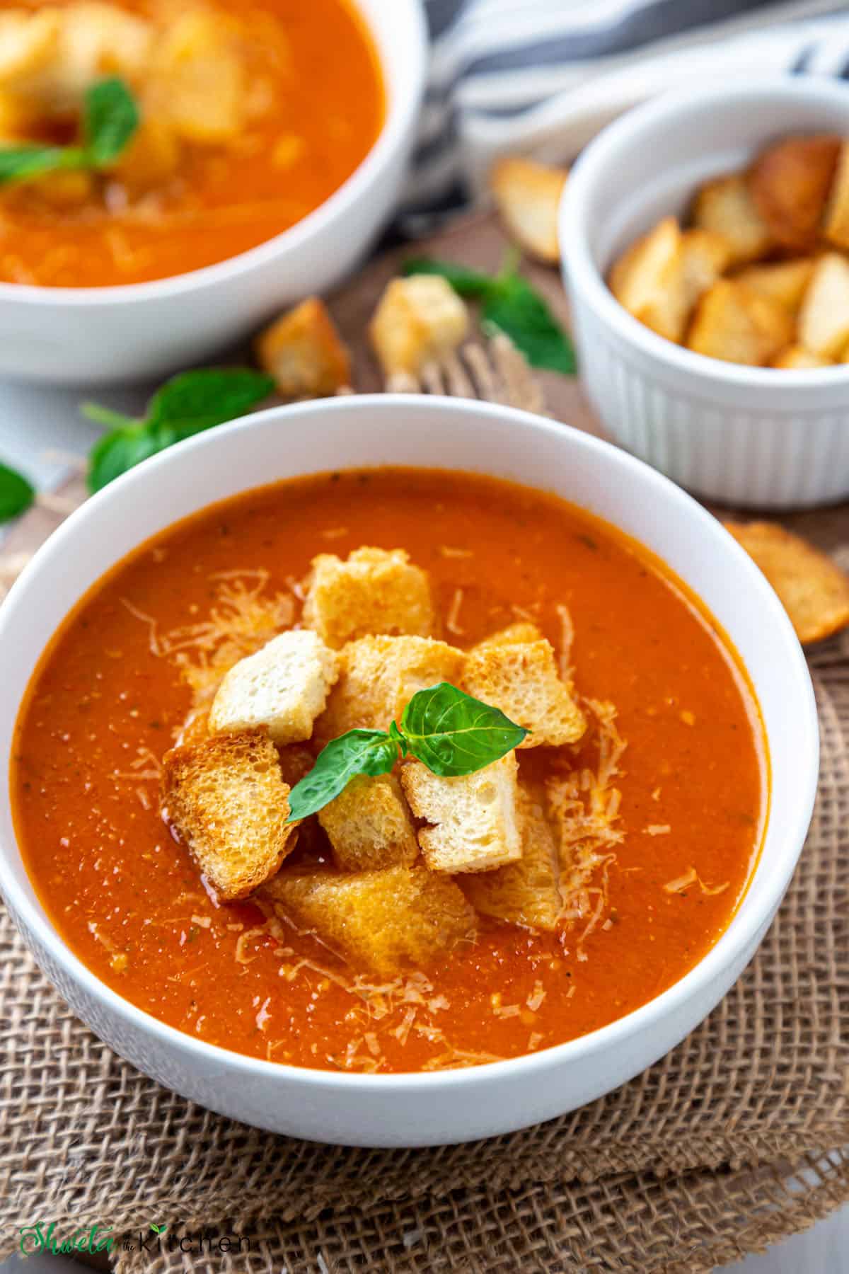 Bowl filled with vegan tomato basil soup topped with croutons and basil placed on burlap cloth. 