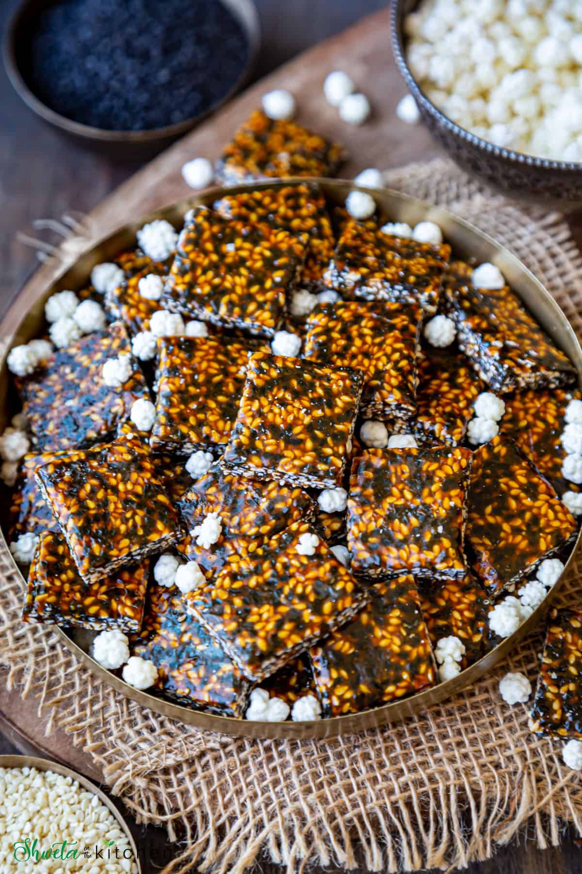 sesame chikki on a plate with sugar coated candy and white and black sesame seeds in background
