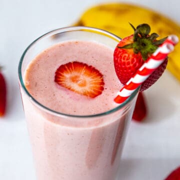 Strawberry banana smoothie in a tall glass with red stripped straw and strawberry on the side and a sliced one at the centre