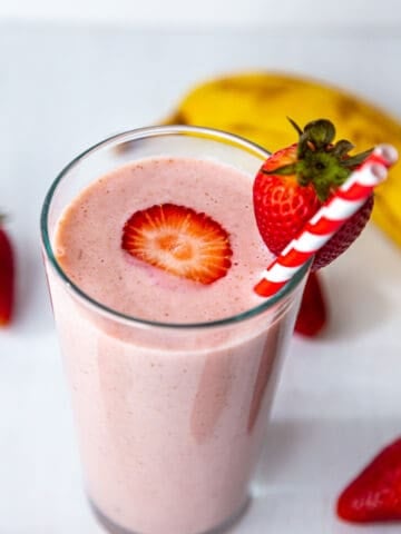 Strawberry banana smoothie in a tall glass with red stripped straw and strawberry on the side and a sliced one at the centre