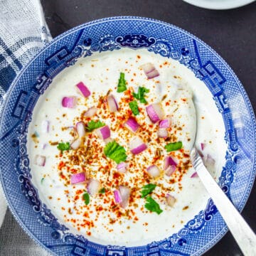 Onion raita served in a blue and white bowl with a spoon and garnished with spices and fresh herbs