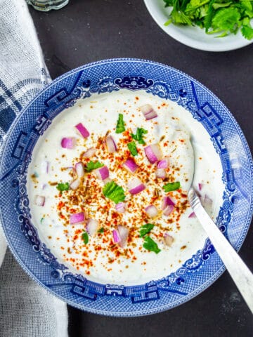 Onion raita served in a blue and white bowl with a spoon and garnished with spices and fresh herbs