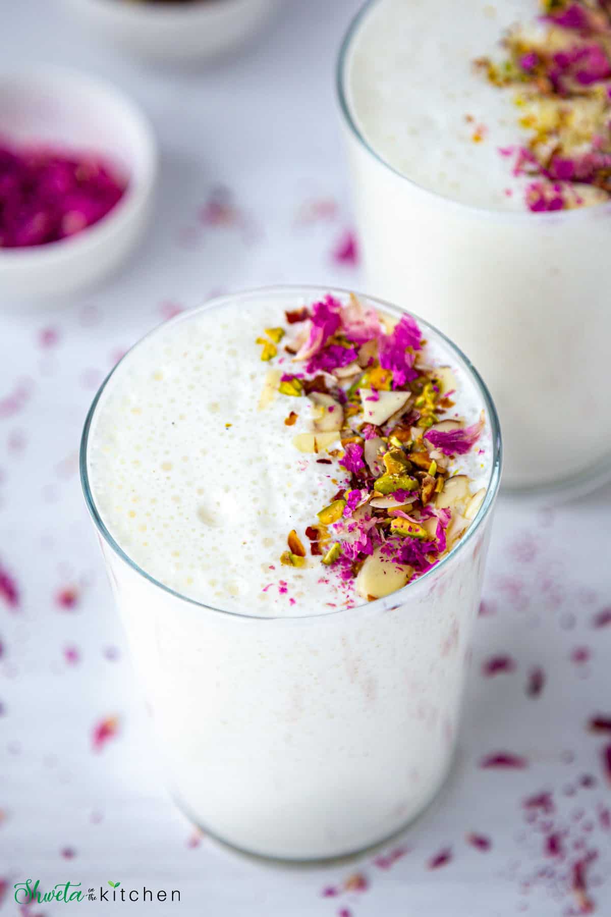 Side angled view of two glasses full of sweet lassi garnished with nuts and rose petals