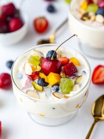 Two bowls of Indian fruit salad garnished with more fruits topped with cherry and golden spoon