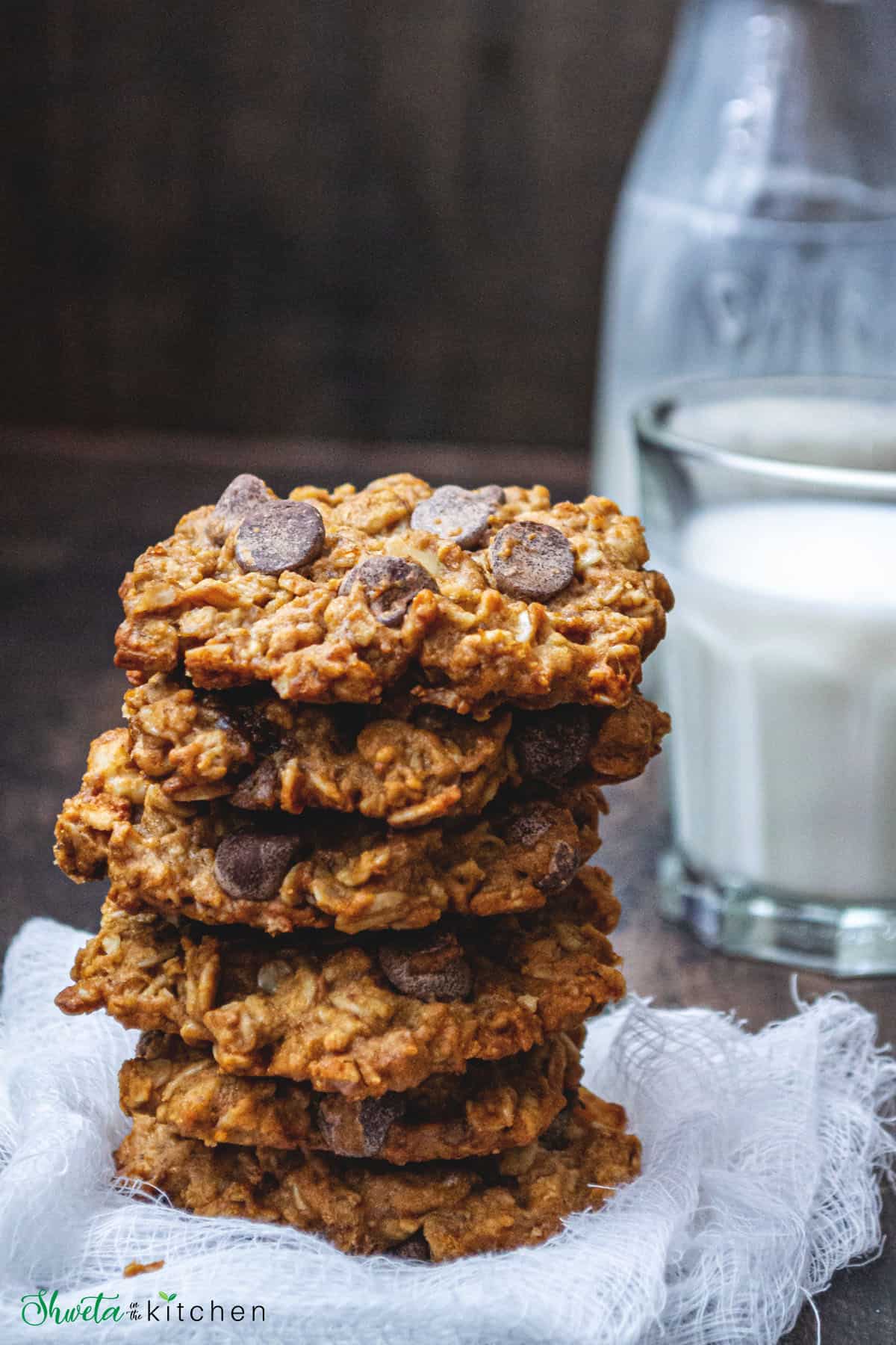 Six chocolate chip oatmeal cookies stacked on top of each other in front of a glass of milk