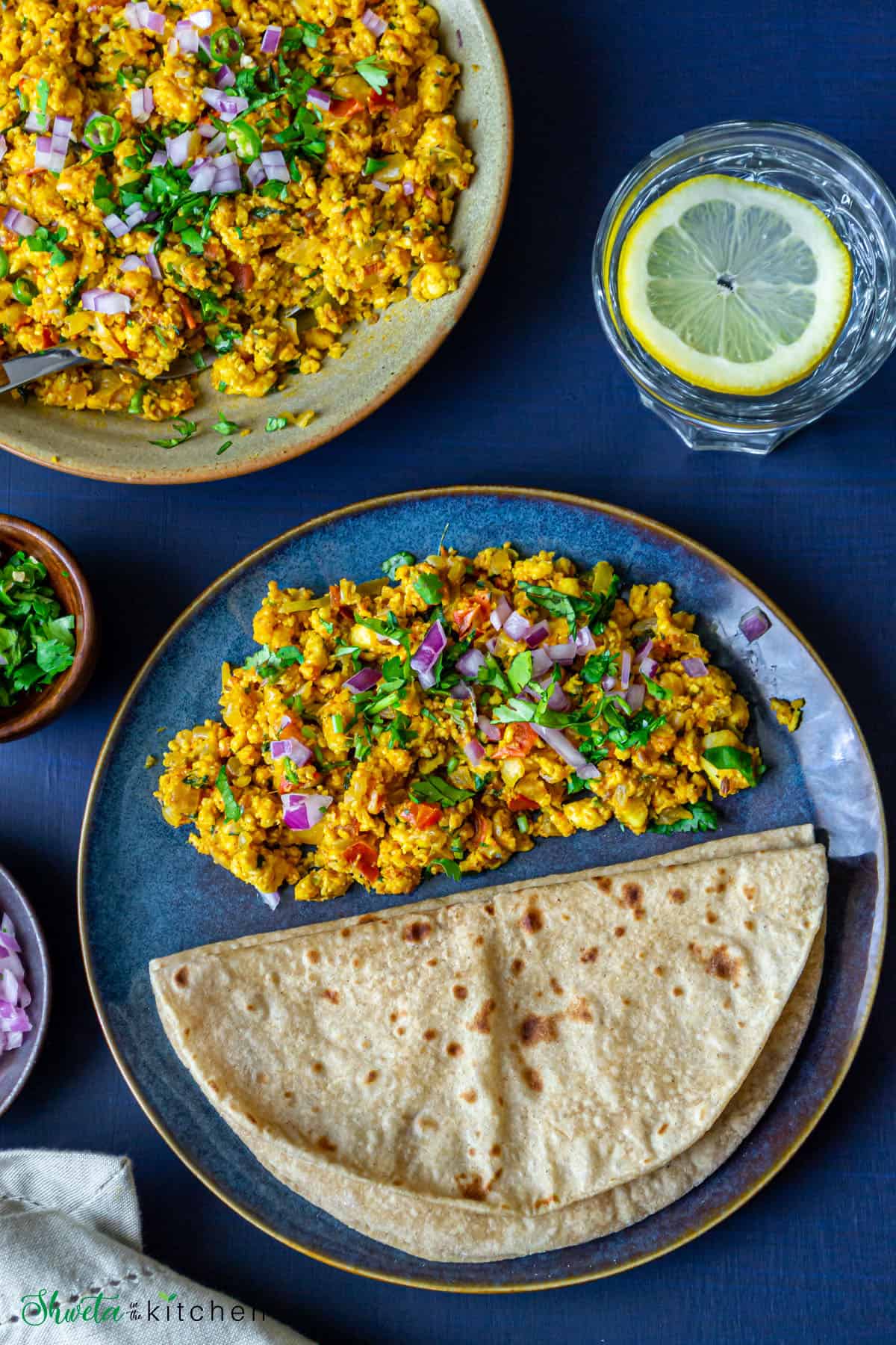 Paneer bhurji served on a blue plate with roti next to a glass of water with a slice of lemon