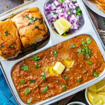 Pav bhaji served on steel plate topped with butter and cilantro with pav, onions and lemon on side