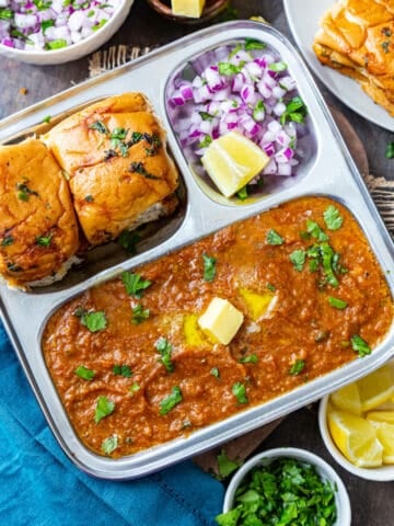 Pav bhaji served on steel plate topped with butter and cilantro with pav, onions and lemon on side