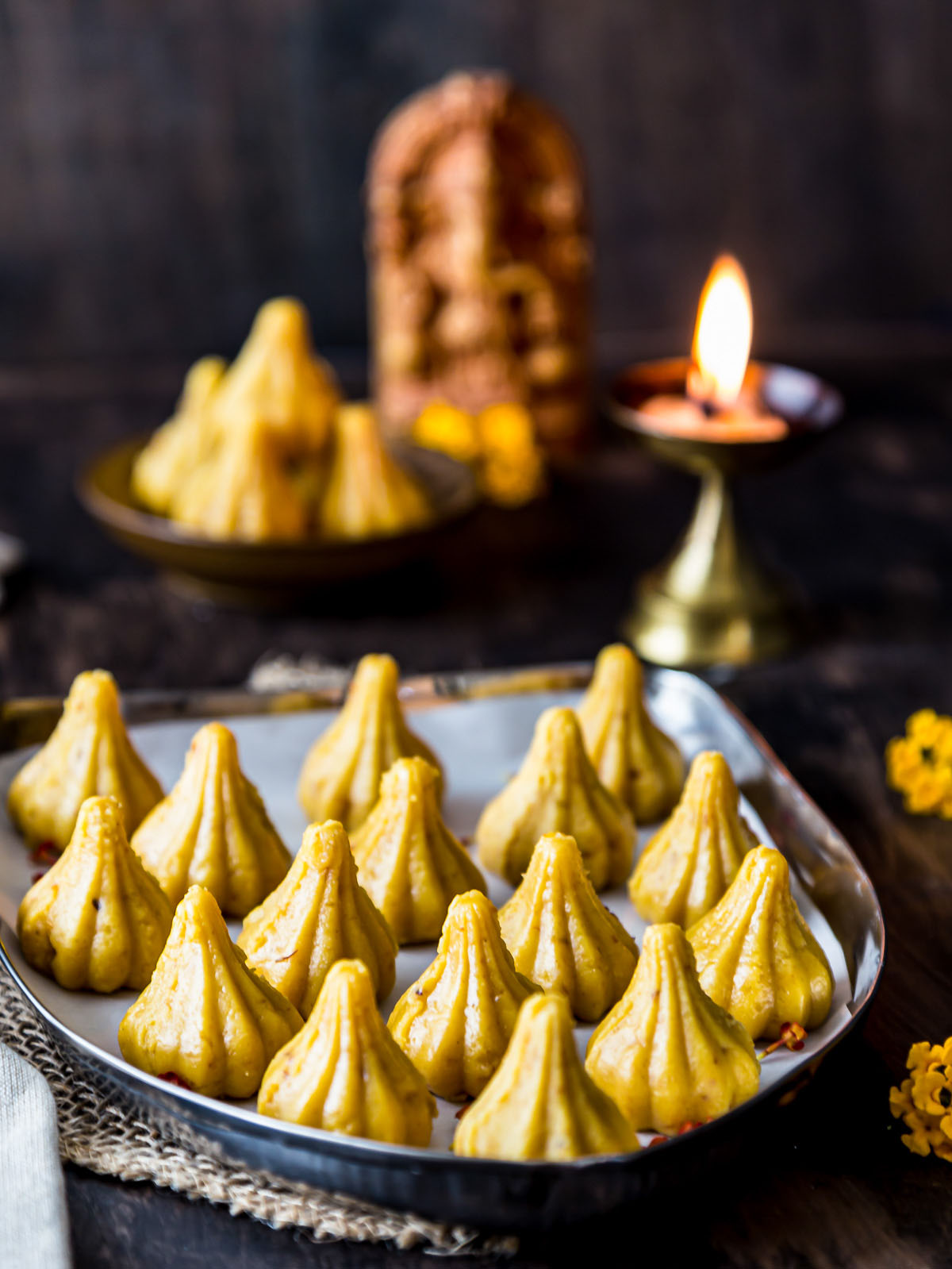 front view of yellow Modak arranged on a plate with light lamp and Ganesh idol behind in background
