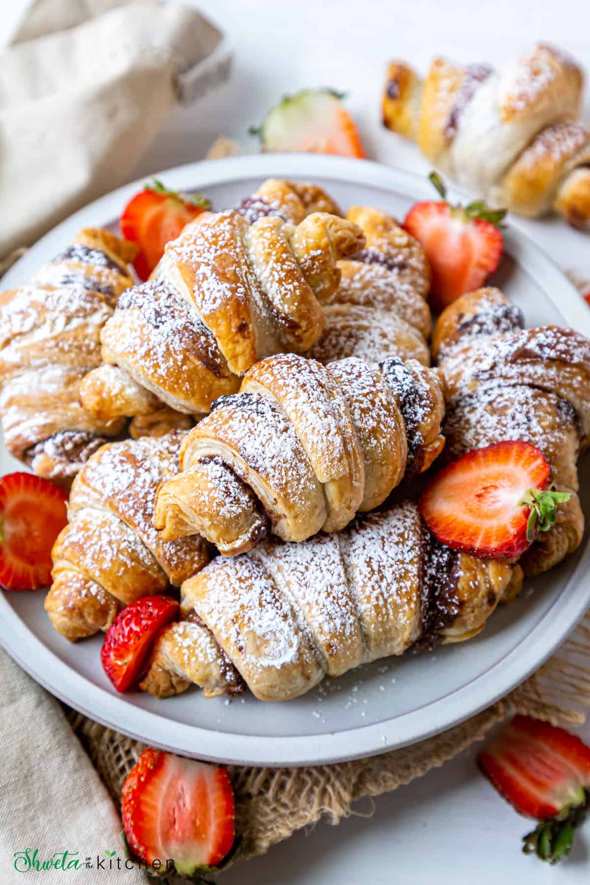 Nutella croissants dusted with powdered sugar piled on a plate along with cut strawberries