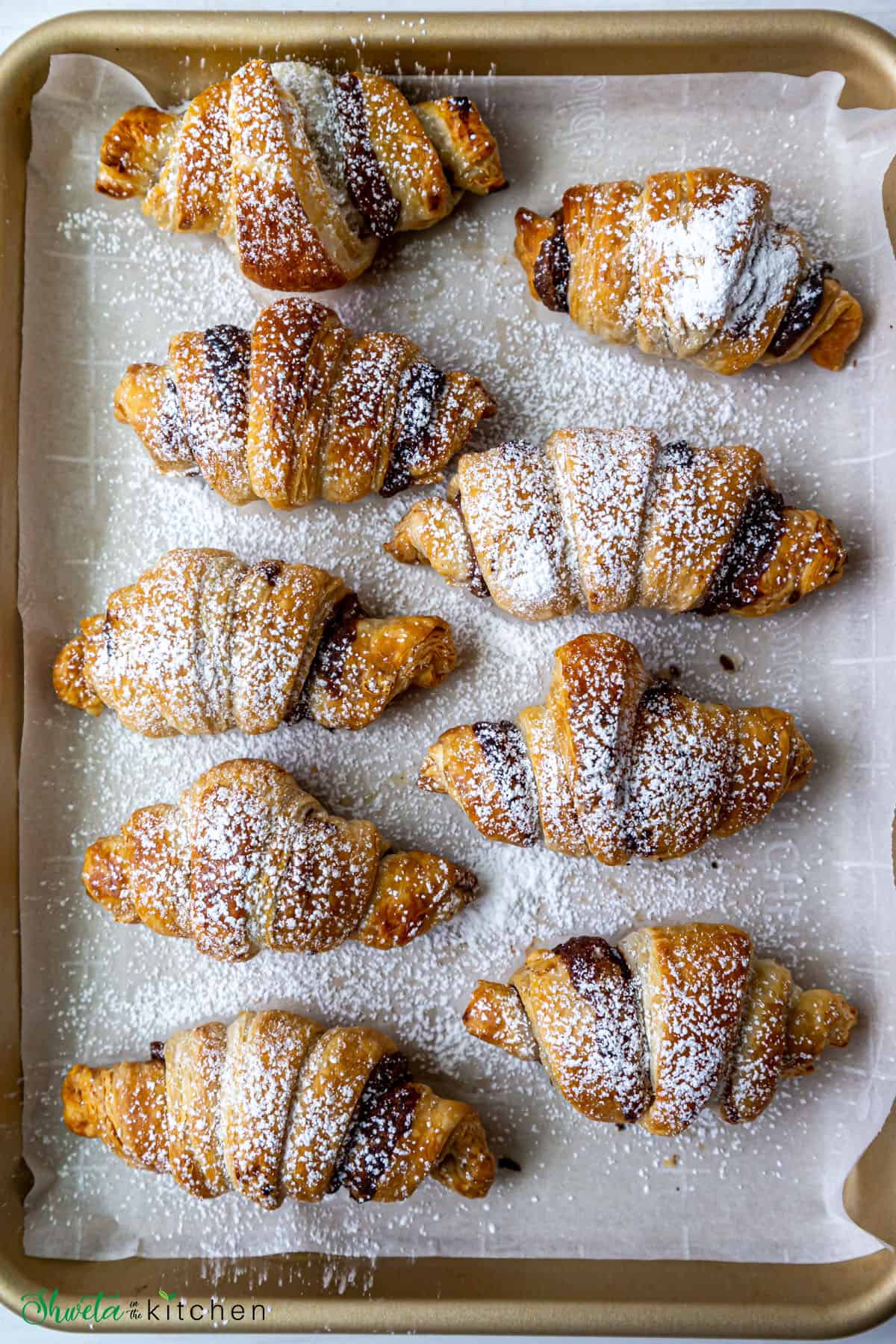 9 Nutella croissants dusted with powdered sugar on a lined baking sheet