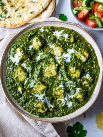 Bowl of palak paneer drizzled with cream in center with plate of naan and salad bowl on side