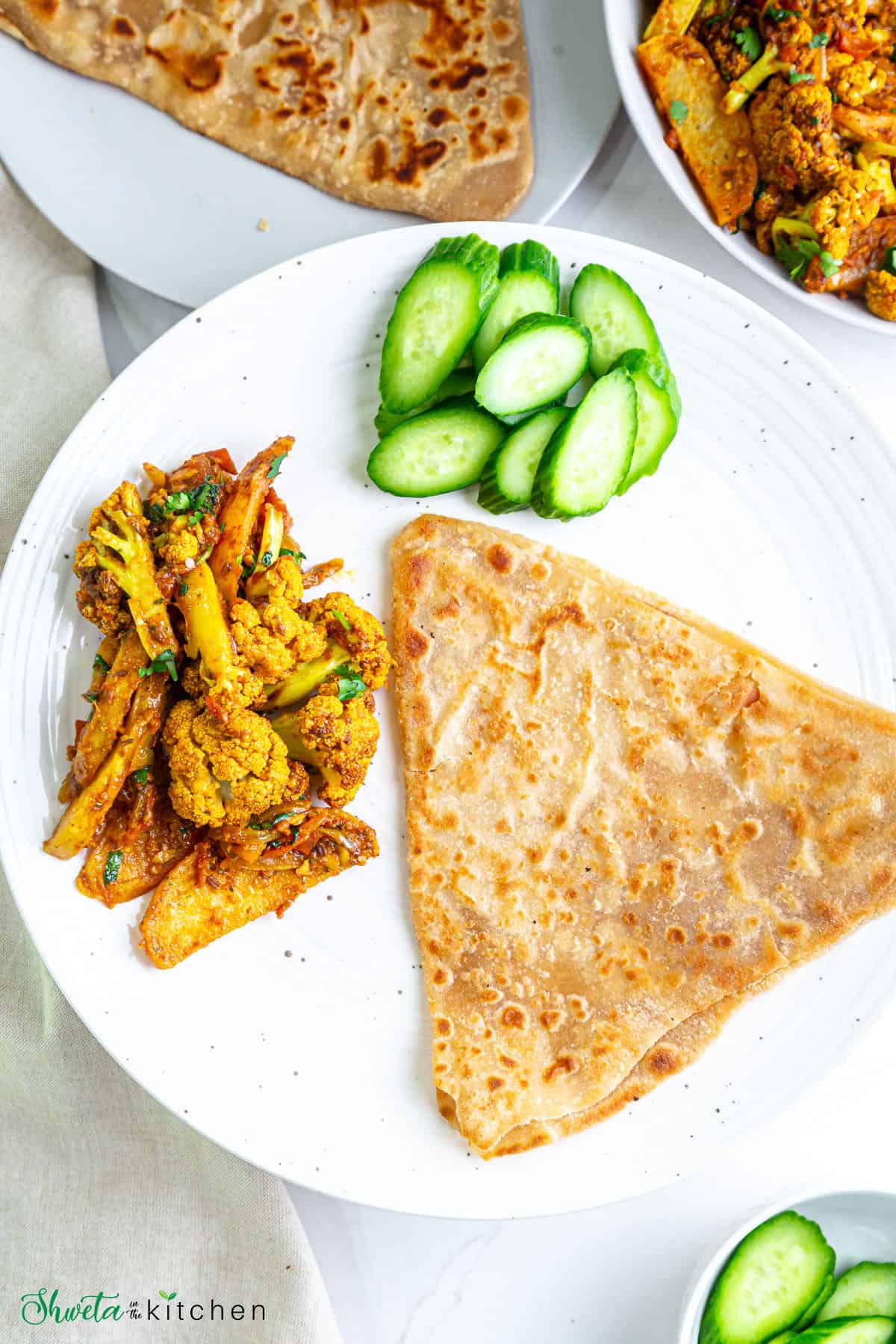 Aloo gobi curry on a plate with paratha and sliced cucumbers