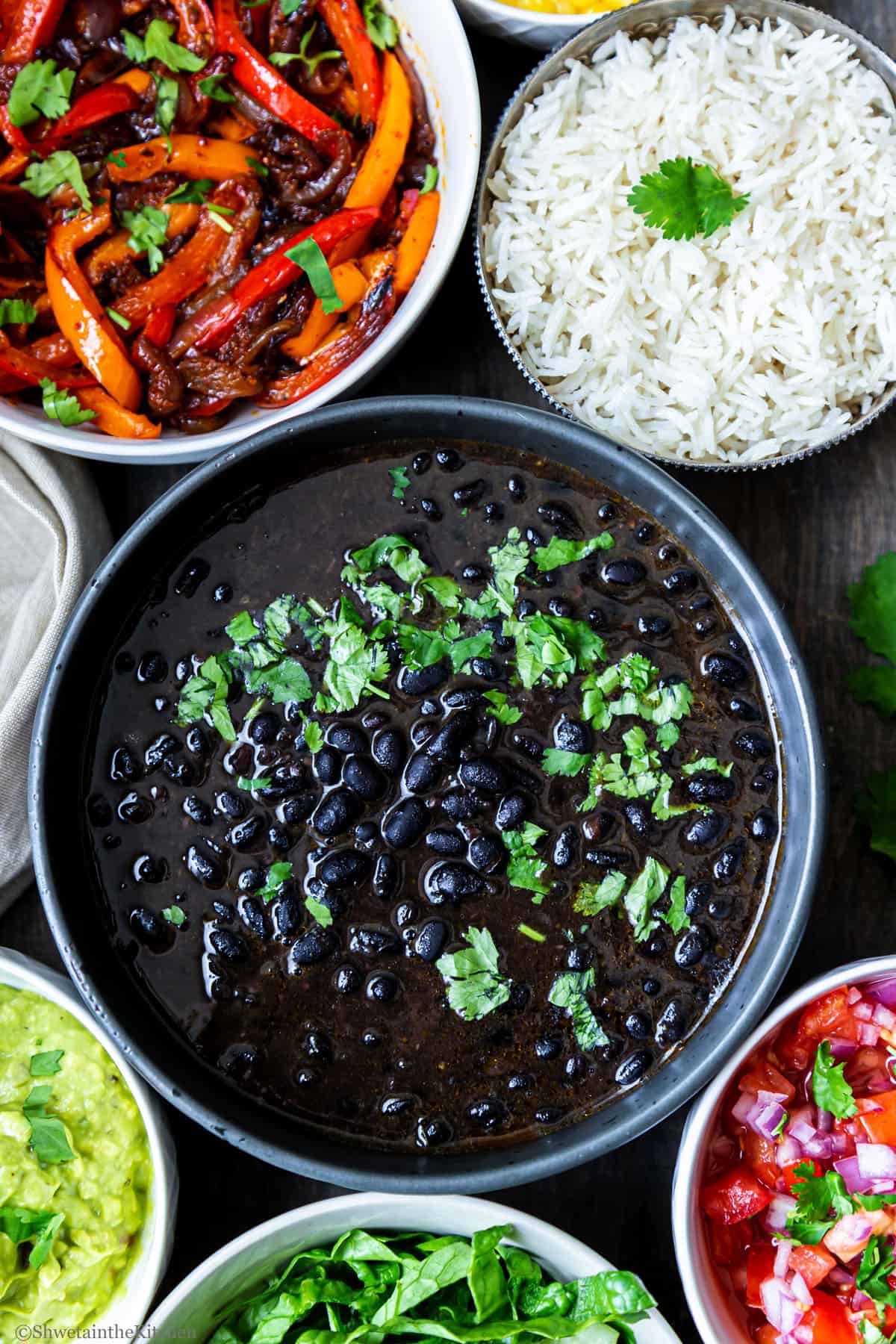 Top view of Instant Pot black beans curry bowl in center with bowls of rice, pico, guacamole, lettuce, and fajita veggies around.
